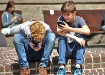 Due adolescenti seduti su un muretto che chiacchierano guardando il cellulare