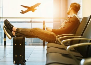 Uomo seduto in aeroporto mentre aspetta al gate. Ue, nuove regole su bagagli a mano: più trasparenza e tariffe standard per i voli