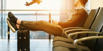 Uomo seduto in aeroporto mentre aspetta al gate. Ue, nuove regole su bagagli a mano: più trasparenza e tariffe standard per i voli