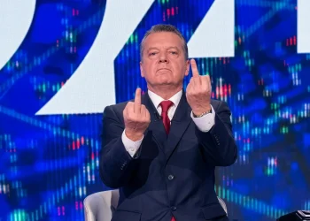 Uomo in giacca e cravatta mostra i diti medi con espressione seria.