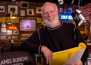 James Burrows sorride, seduto accanto alla sua sedia da regista, con copione.