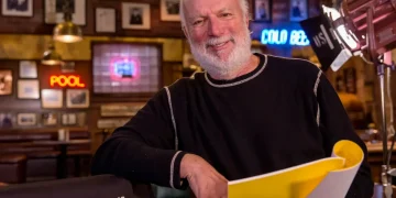 James Burrows sorride, seduto accanto alla sua sedia da regista, con copione.