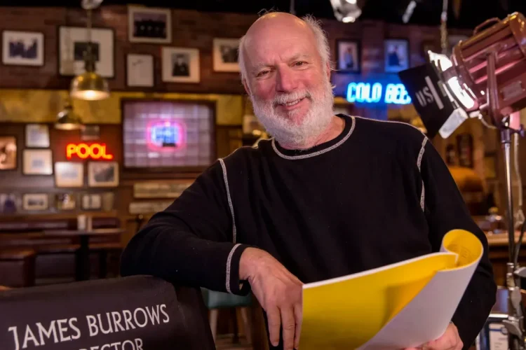 James Burrows sorride, seduto accanto alla sua sedia da regista, con copione.