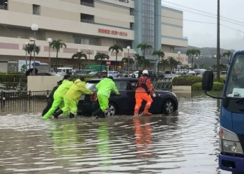 Persone in impermeabili spingono auto allagata durante alluvione in Giappone.