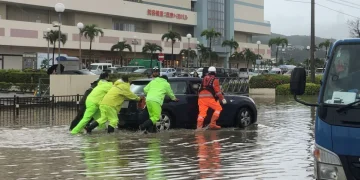 Persone in impermeabili spingono auto allagata durante alluvione in Giappone.