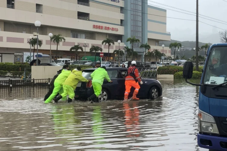 Persone in impermeabili spingono auto allagata durante alluvione in Giappone.