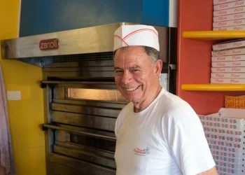 Pizzaiolo sorridente in pizzeria davanti al forno e scatole pizza.