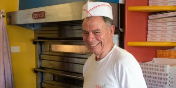 Pizzaiolo sorridente in pizzeria davanti al forno e scatole pizza.