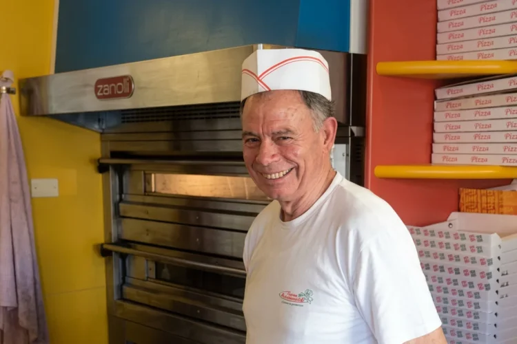 Pizzaiolo sorridente in pizzeria davanti al forno e scatole pizza.