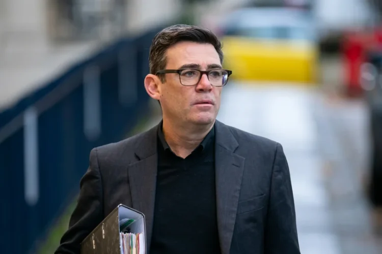 Andy Burnham con occhiali cammina all'aperto portando dei documenti