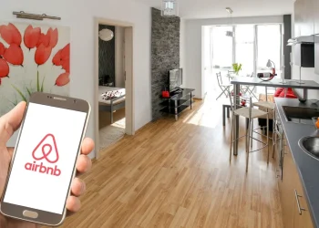 Mano con smartphone mostra app Airbnb in appartamento moderno, per mettere in guardia dalle truffe estive.