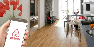 Mano con smartphone mostra app Airbnb in appartamento moderno, per mettere in guardia dalle truffe estive.