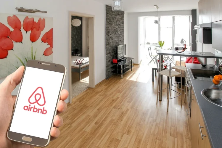 Mano con smartphone mostra app Airbnb in appartamento moderno, per mettere in guardia dalle truffe estive.