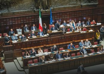 Deputati siedono nell'aula parlamentare italiana con bandiere visibili, focus sull'assemblea. Riforma elettorale, ok dalla Camera con 217 sì: il testo va al Senato tra le tensioni di governo