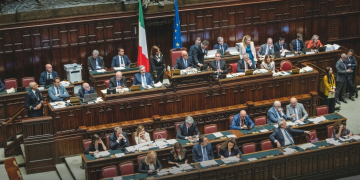 Deputati siedono nell'aula parlamentare italiana con bandiere visibili, focus sull'assemblea. Riforma elettorale, ok dalla Camera con 217 sì: il testo va al Senato tra le tensioni di governo