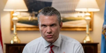 Pete Hegseth punta il dito, cravatta rossa, bandiere americane sullo sfondo