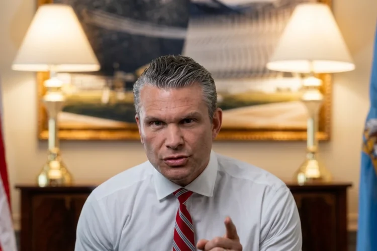 Pete Hegseth punta il dito, cravatta rossa, bandiere americane sullo sfondo