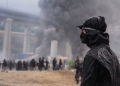 Manifestante con maschera e cappuccio nero osserva fumo in Val di Susa. Scontri No Tav in Val di Susa: 450 manifestanti identificati e oltre 120 feriti tra le forze dell'ordine
