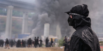 Manifestante con maschera e cappuccio nero osserva fumo in Val di Susa. Scontri No Tav in Val di Susa: 450 manifestanti identificati e oltre 120 feriti tra le forze dell'ordine