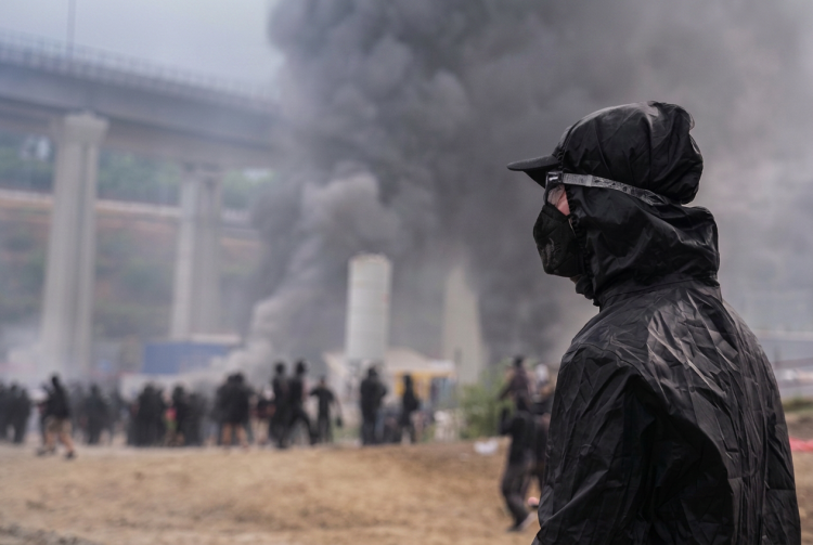 Manifestante con maschera e cappuccio nero osserva fumo in Val di Susa. Scontri No Tav in Val di Susa: 450 manifestanti identificati e oltre 120 feriti tra le forze dell'ordine