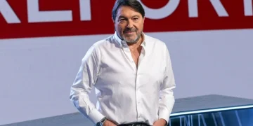 Sigfrido Ranucci sorride in studio con logo Report rosso.
