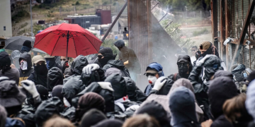 Folla di manifestanti protesta con passamontagna vicino a barriera metallica. Fermati quattro stranieri per violenze ai cantieri Tav, ipotesi devastazione al vaglio