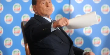 Silvio Berlusconi seduto con espressione seria durante un intervento pubblico