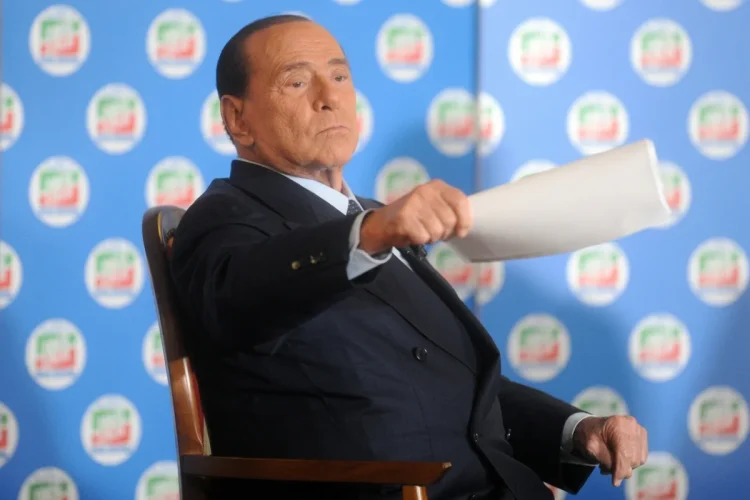 Silvio Berlusconi seduto con espressione seria durante un intervento pubblico
