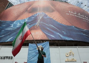 an-iranian-man-holds-iran-s-national-flag-in-front-of-a-huge-anti-us-billboard-r-1200x800.webp