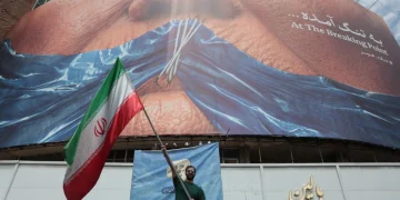 an-iranian-man-holds-iran-s-national-flag-in-front-of-a-huge-anti-us-billboard-r-1200x800.webp