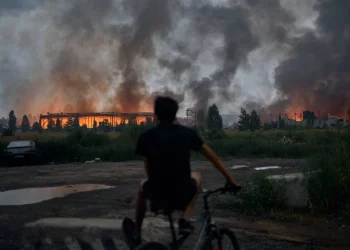 Uomo in bicicletta osserva edifici distrutti avvolti dalle fiamme e fumo