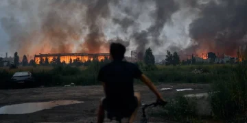 Uomo in bicicletta osserva edifici distrutti avvolti dalle fiamme e fumo