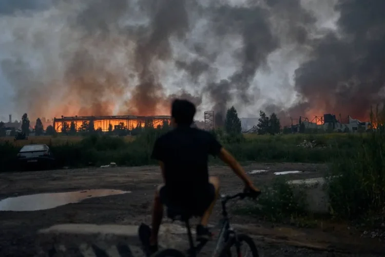 Uomo in bicicletta osserva edifici distrutti avvolti dalle fiamme e fumo