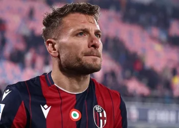 Calciatore maglia rossoblù applaude in campo, sguardo pensieroso.