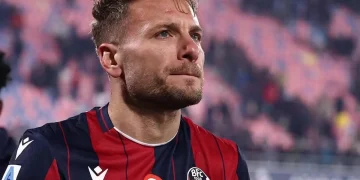 Calciatore maglia rossoblù applaude in campo, sguardo pensieroso.