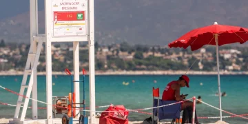 Bagnino in divisa rossa seduto in spiaggia con telefono, torretta di salvataggio.