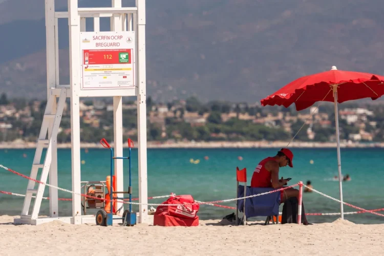 Bagnino in divisa rossa seduto in spiaggia con telefono, torretta di salvataggio.