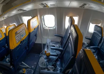 Interno aereo Ryanair danneggiato, finestrino mancante, detriti, maschere ossigeno pendenti.