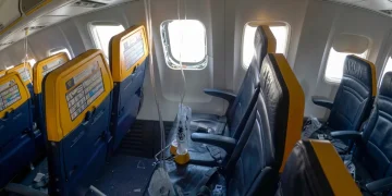 Interno aereo Ryanair danneggiato, finestrino mancante, detriti, maschere ossigeno pendenti.