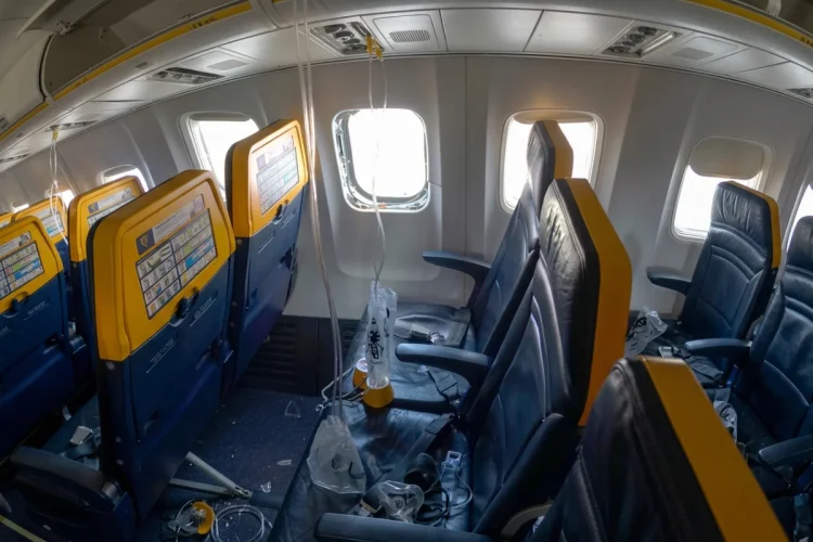 Interno aereo Ryanair danneggiato, finestrino mancante, detriti, maschere ossigeno pendenti.