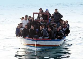 Uomini migranti sovraffollano un piccolo barcone bianco e blu in mare aperto.