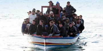 Uomini migranti sovraffollano un piccolo barcone bianco e blu in mare aperto.