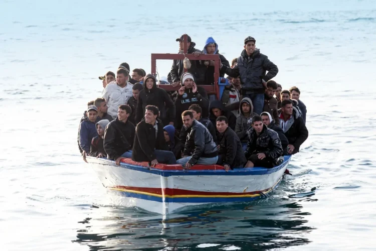 Uomini migranti sovraffollano un piccolo barcone bianco e blu in mare aperto.