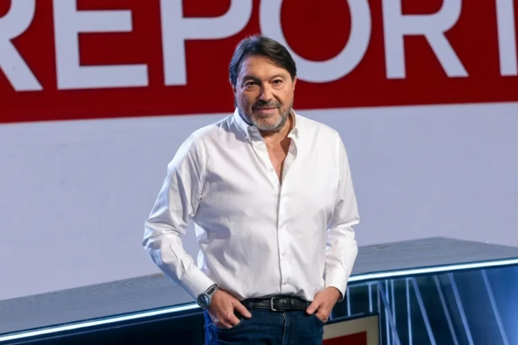 Sigfrido Ranucci sorride in studio con logo Report rosso.
