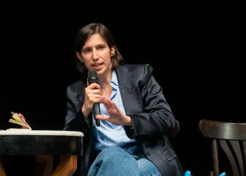 Elly Schlein parla al microfono gesticolando, indossa giacca scura.