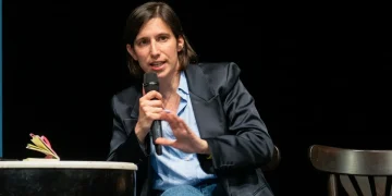 Elly Schlein parla al microfono gesticolando, indossa giacca scura.