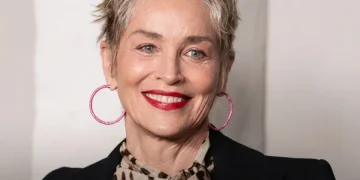 Sharon Stone sorride con orecchini rosa, capelli corti e giacca scura.