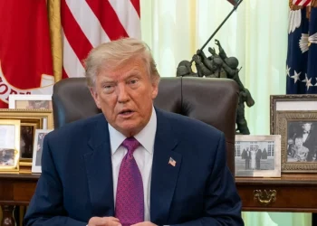 Donald Trump siede al tavolo, bandiere americane e foto dietro.