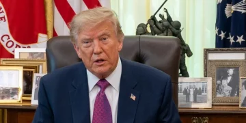 Donald Trump siede al tavolo, bandiere americane e foto dietro.