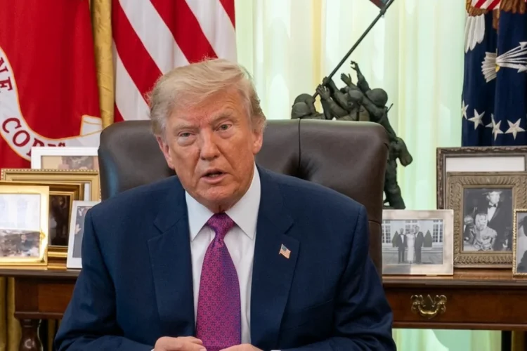 Donald Trump siede al tavolo, bandiere americane e foto dietro.